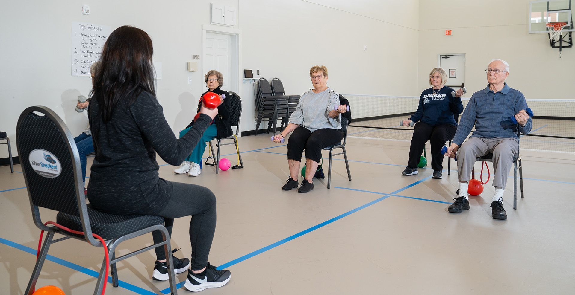 An instructor leads a group of adults in a seated exercise class with handheld weights in a gym.