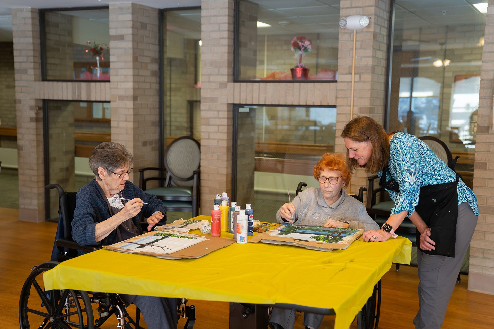 Two people sitting at a table painting on canvases with an instructor standing nearby in a brightly lit community room.