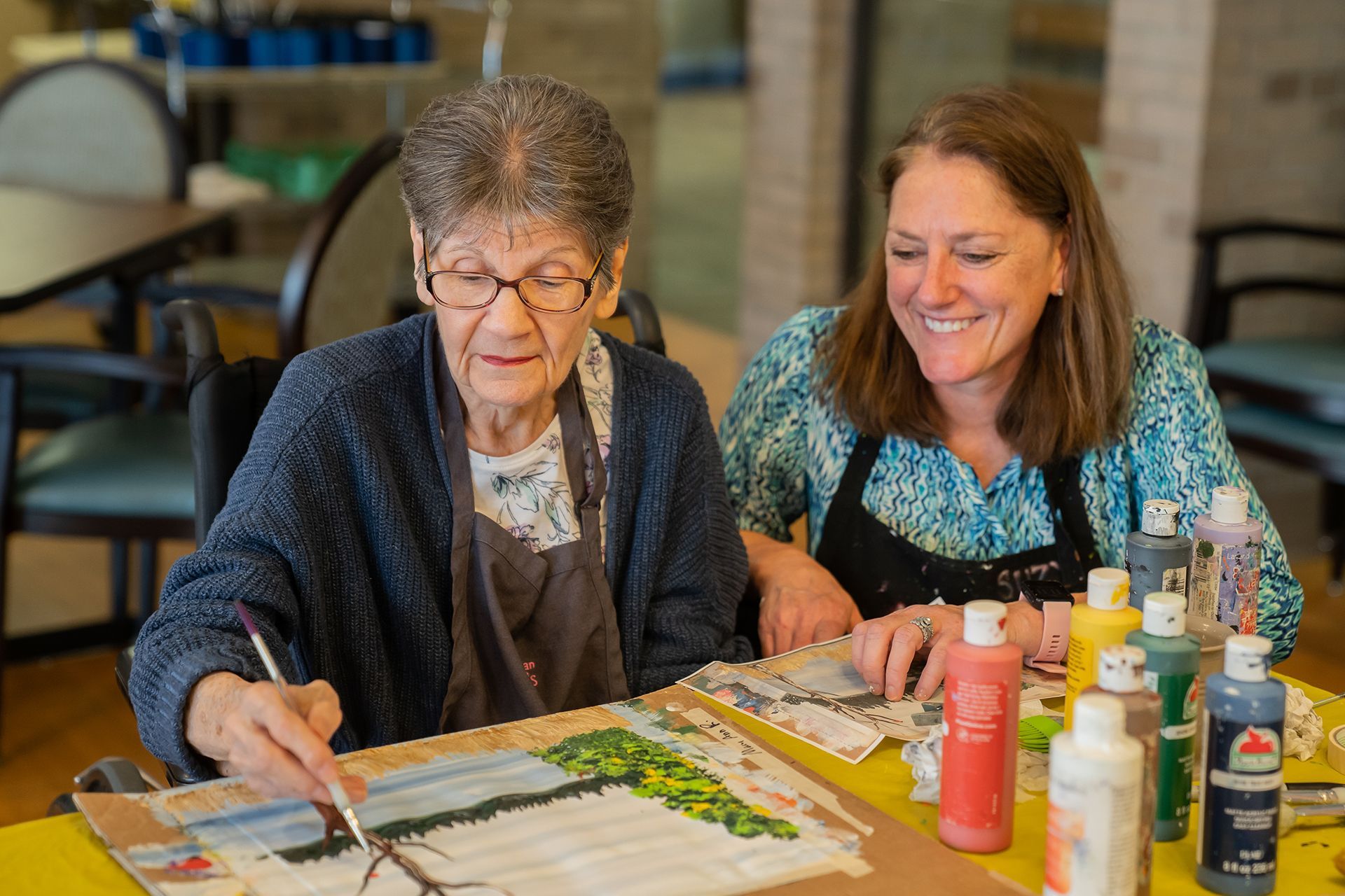 An older adult and a younger companion smile while painting on canvas together at a table with colorful paint bottles.