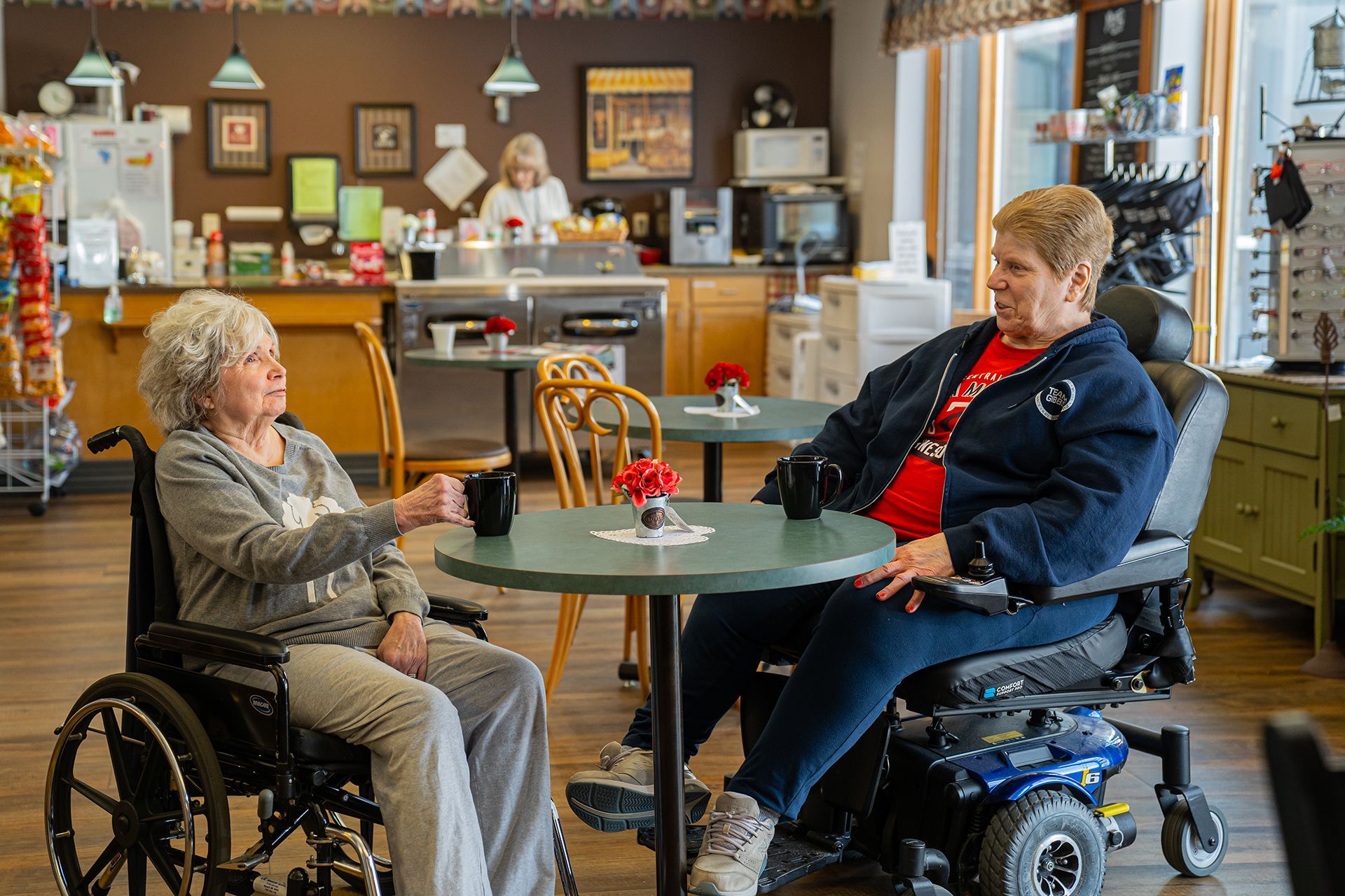 Two people in wheelchairs sit at a table in a cafe, conversing over drinks.