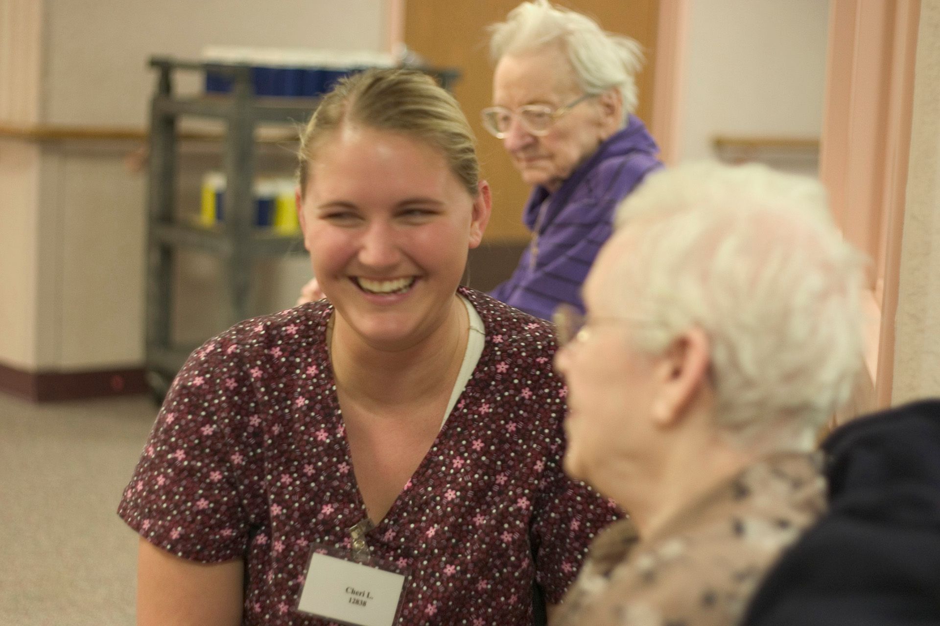 A smiling person wearing a floral top and name tag interacts with an older individual in a community or care setting.