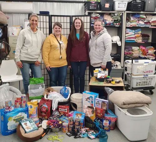 Four people stand in an animal shelter supply room, behind a collection of donated pet food, toys, and supplies.