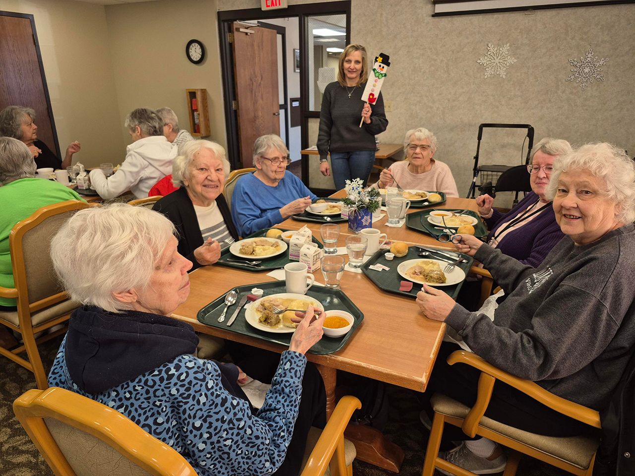 Group of people at a dining table with plates of food, with a person standing in the background holding a decoration.