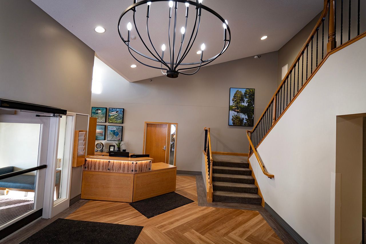A hotel lobby features a wooden reception desk, a chandelier, dark stairs with wooden railings, and herringbone floors.