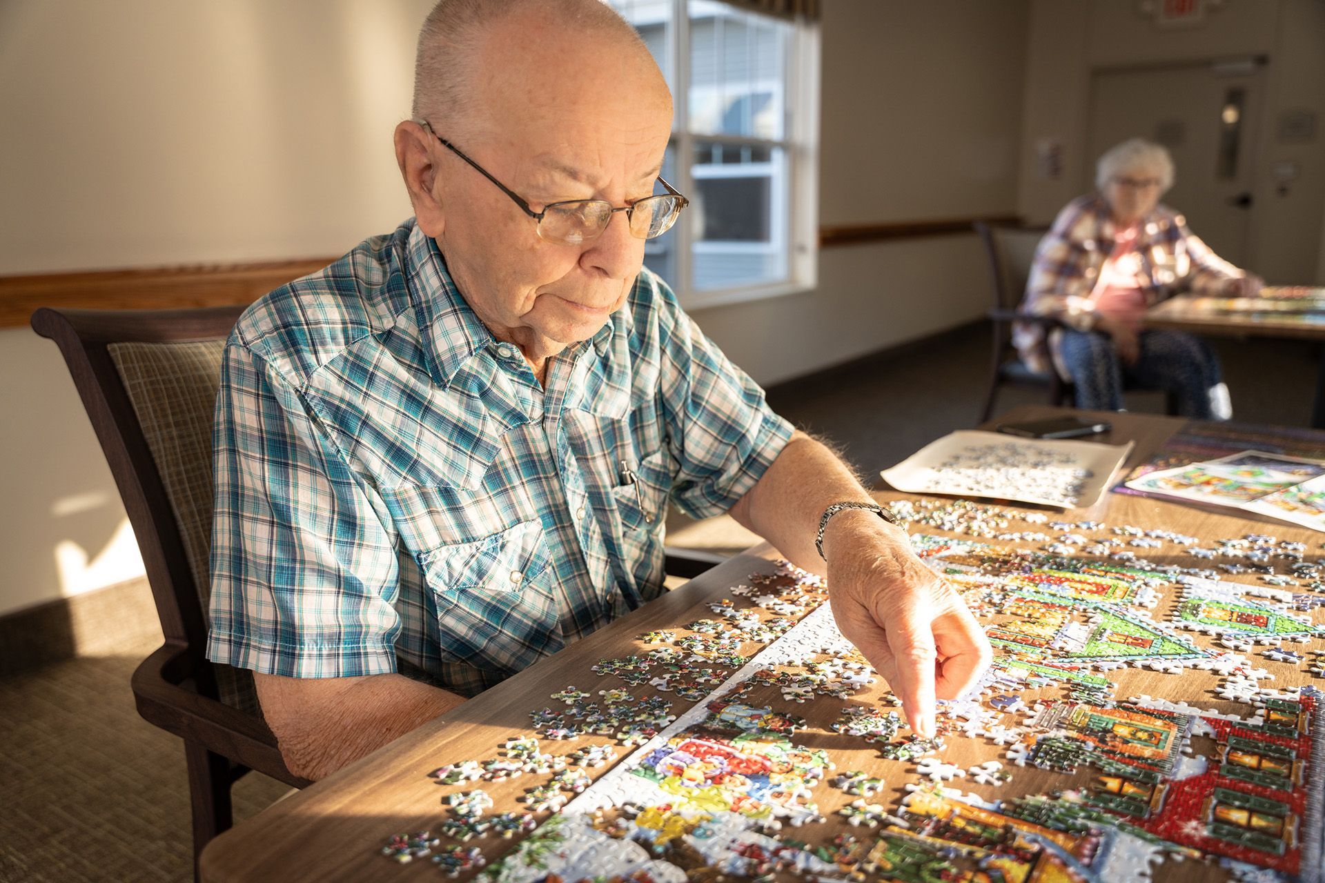 A person in a plaid shirt sits at a table carefully placing a piece into a large, colorful jigsaw puzzle.