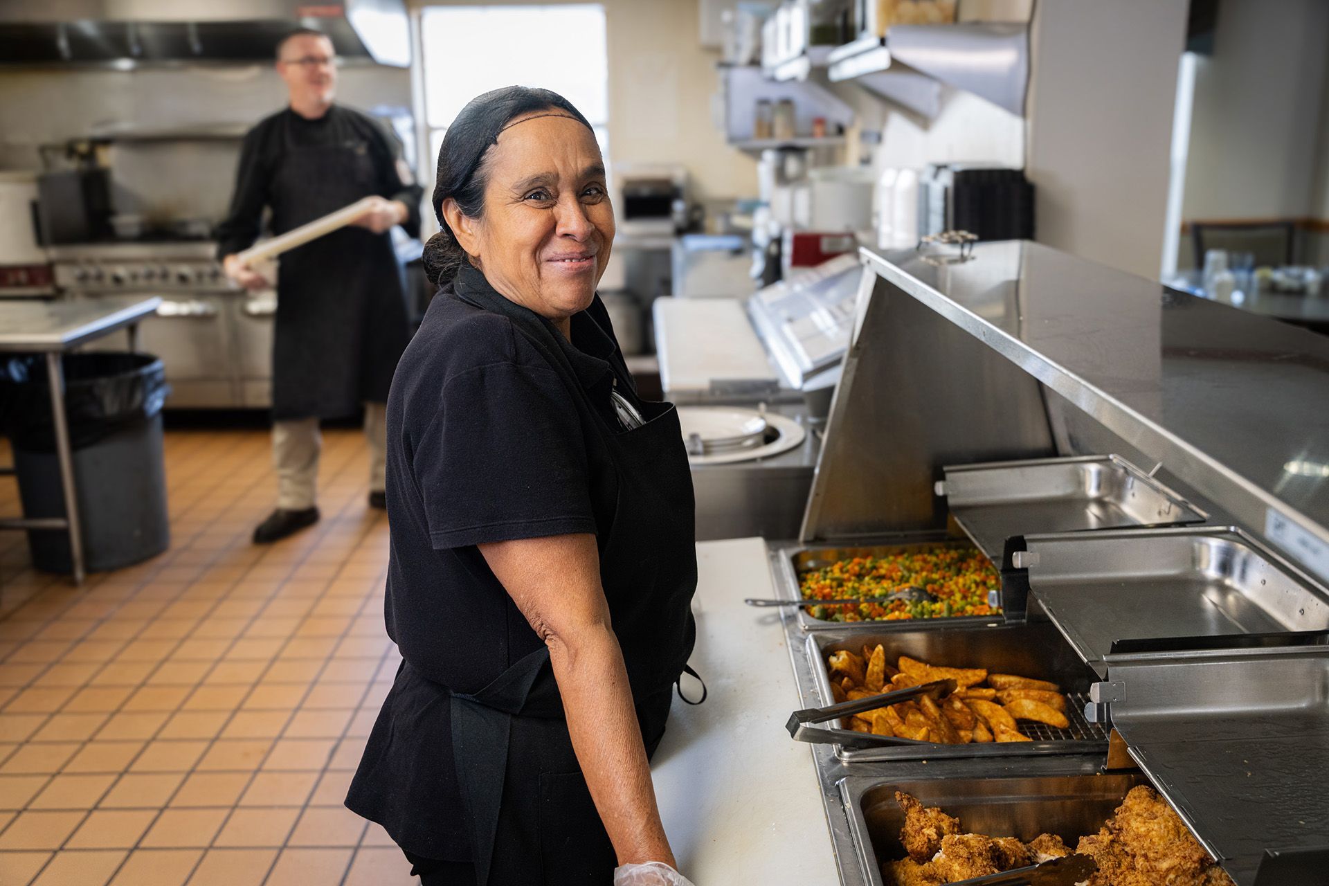 A kitchen employee smiles while standing behind a food warmer, with another staff member working in the background.