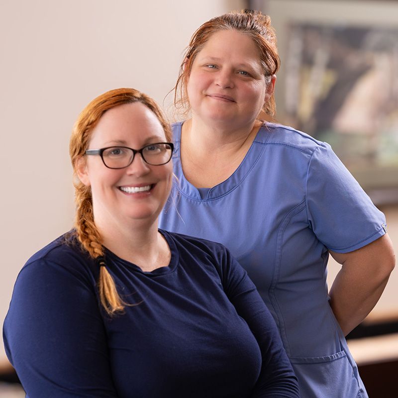 Two smiling women posed together, one in a navy long-sleeve shirt and glasses, the other in light blue medical scrubs.