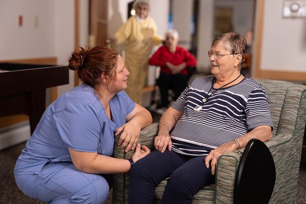 A caregiver kneels beside a resident in a lounge, both engaged in a friendly conversation.