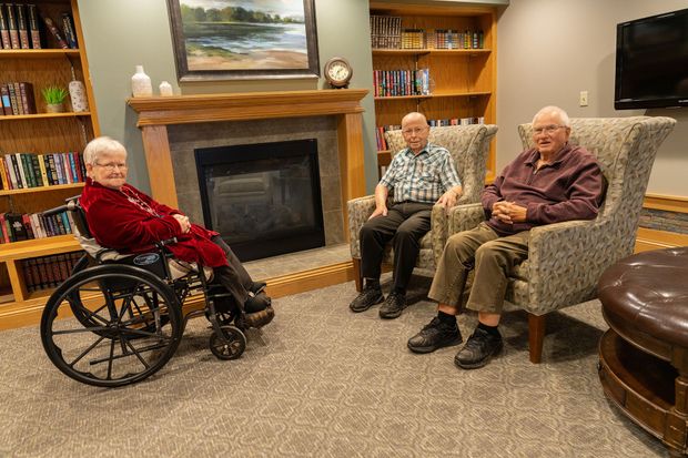 Three people sitting in a cozy room with a fireplace, bookcases, and a television, engaged in conversation.