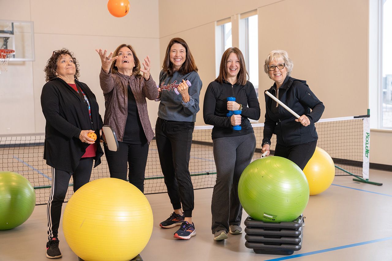 Five people in a gym setting stand near fitness balls, with one tossing an orange ball into the air.