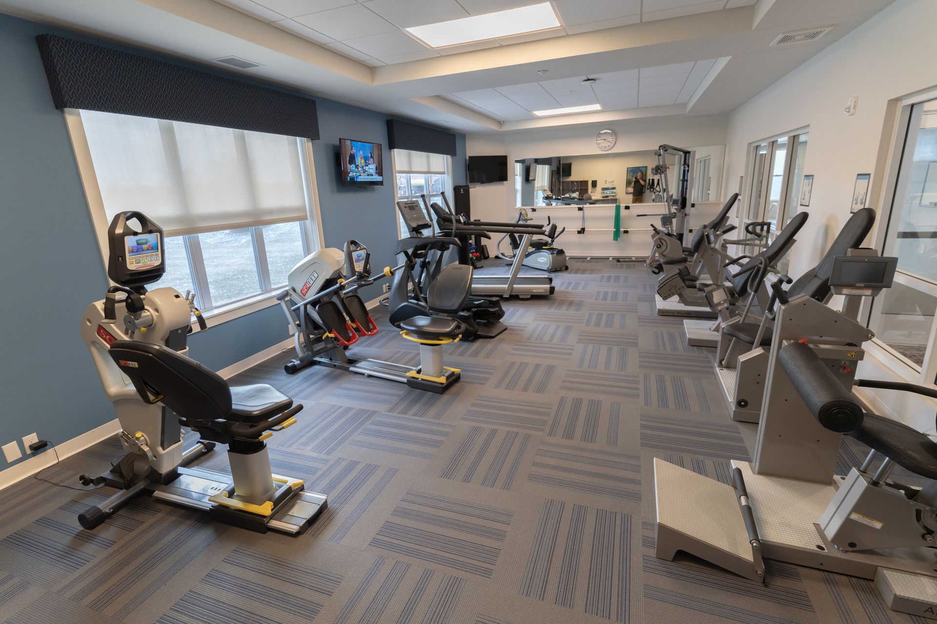 A bright fitness center featuring rows of exercise machines, including stationary bikes and a treadmill on patterned carpet.
