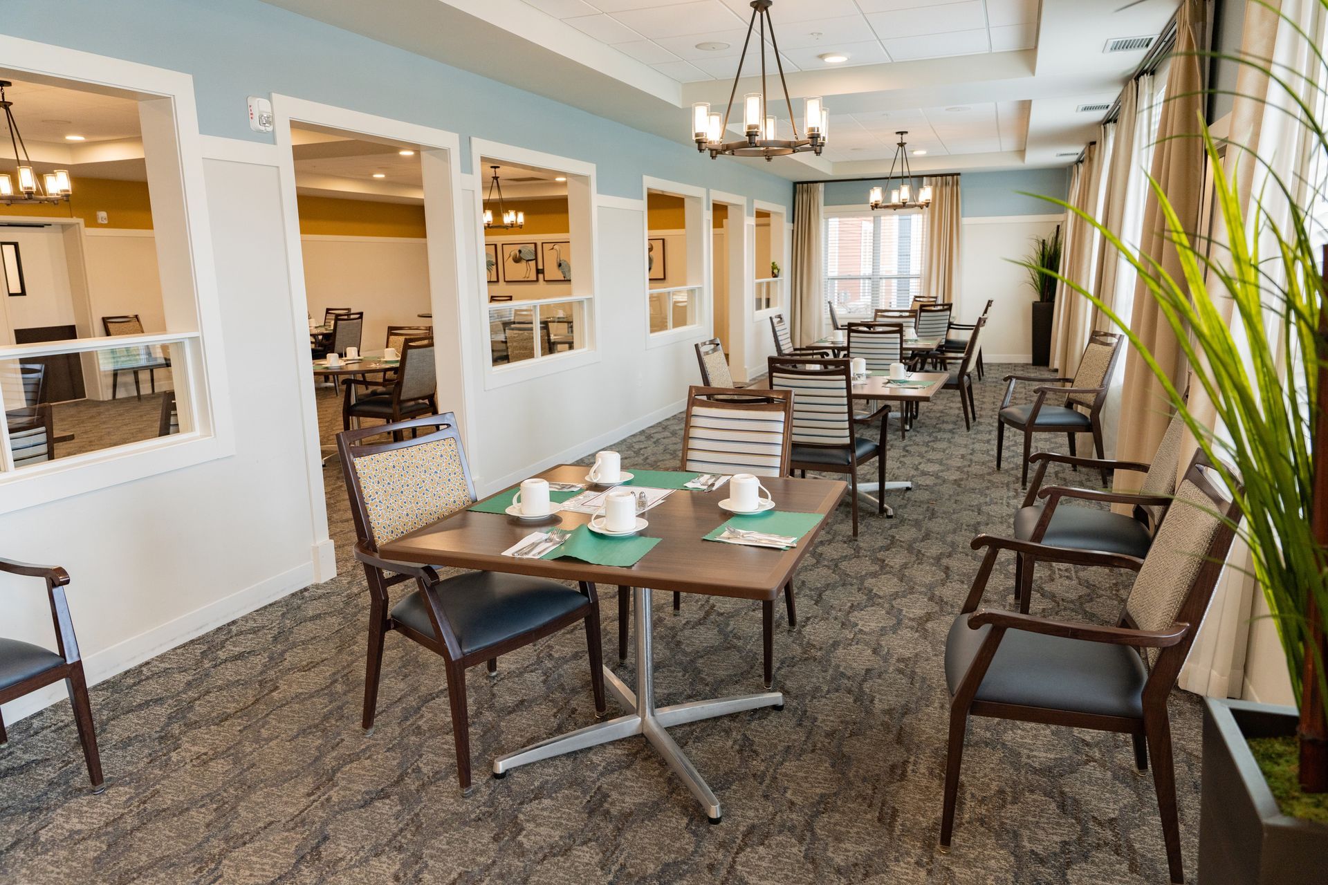A dining room with multiple square tables and chairs, arranged in a row with interior wall cutouts and beige curtains.