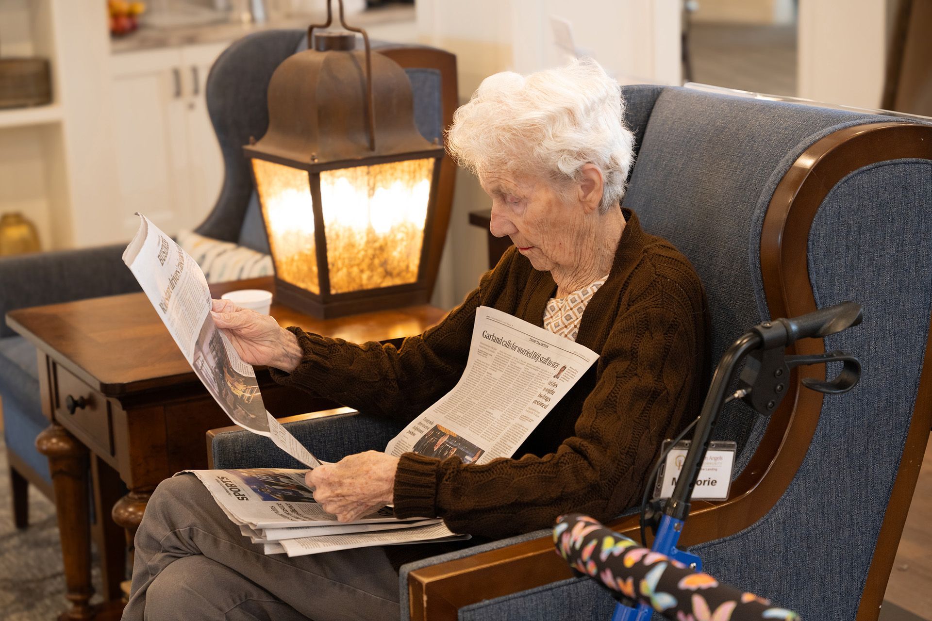 An individual sitting in a blue armchair in a brightly lit room, reading a newspaper with a walker nearby.