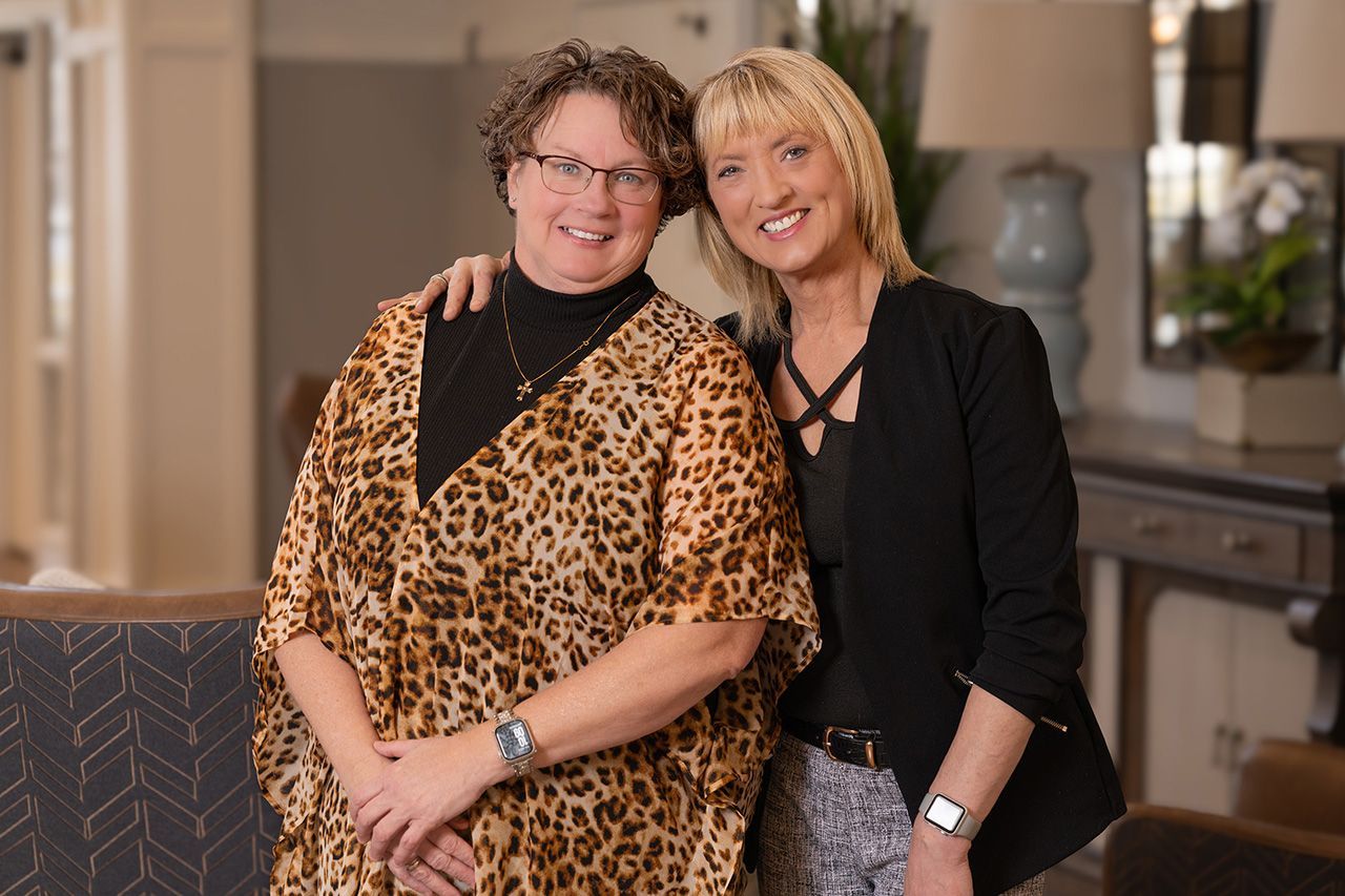 Two people stand side-by-side in an indoor setting, both smiling and looking at the camera.