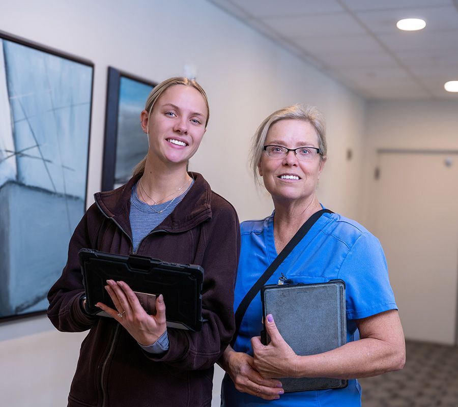 Two people standing in a hallway, both holding tablets and smiling toward the camera.