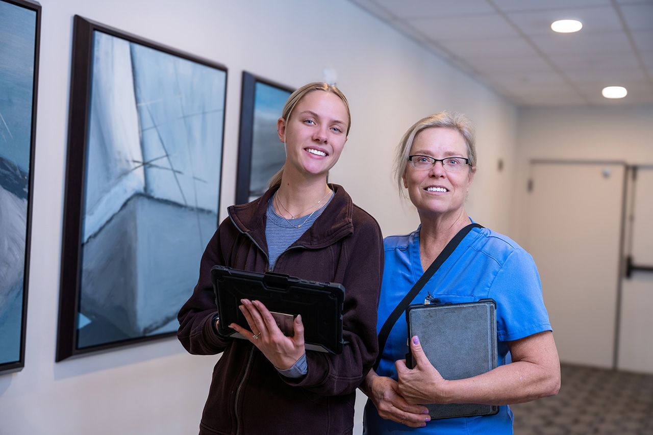 Two people standing in a hallway, one holding a tablet and one a folder, both smiling toward the camera.