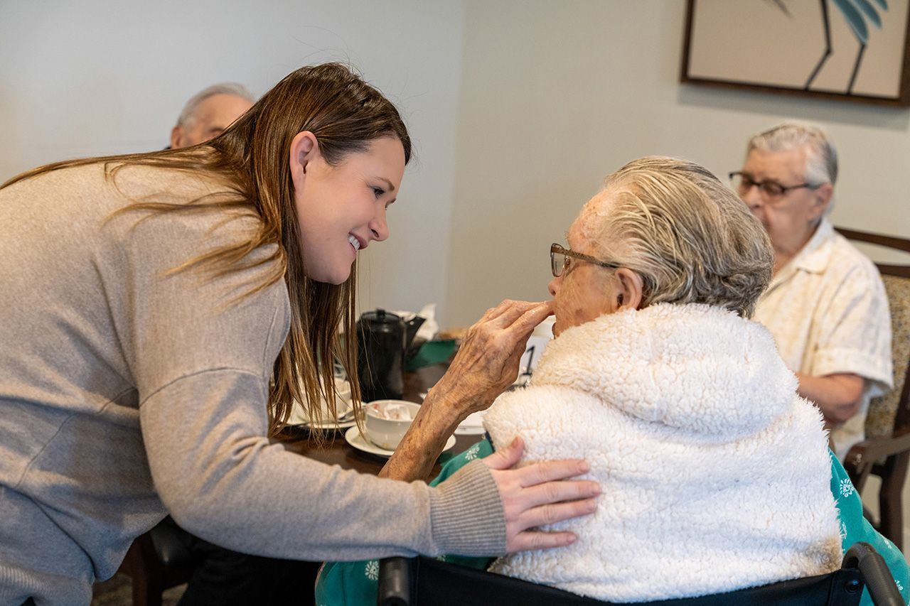 A person in a beige sweater tenderly assists a seated individual with a meal in a dining room setting.