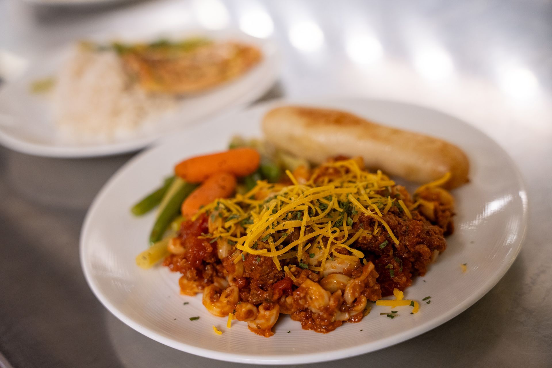 A white plate holds a serving of cheesy chili macaroni, green beans, carrots, and a breadstick. Another dish is blurred.
