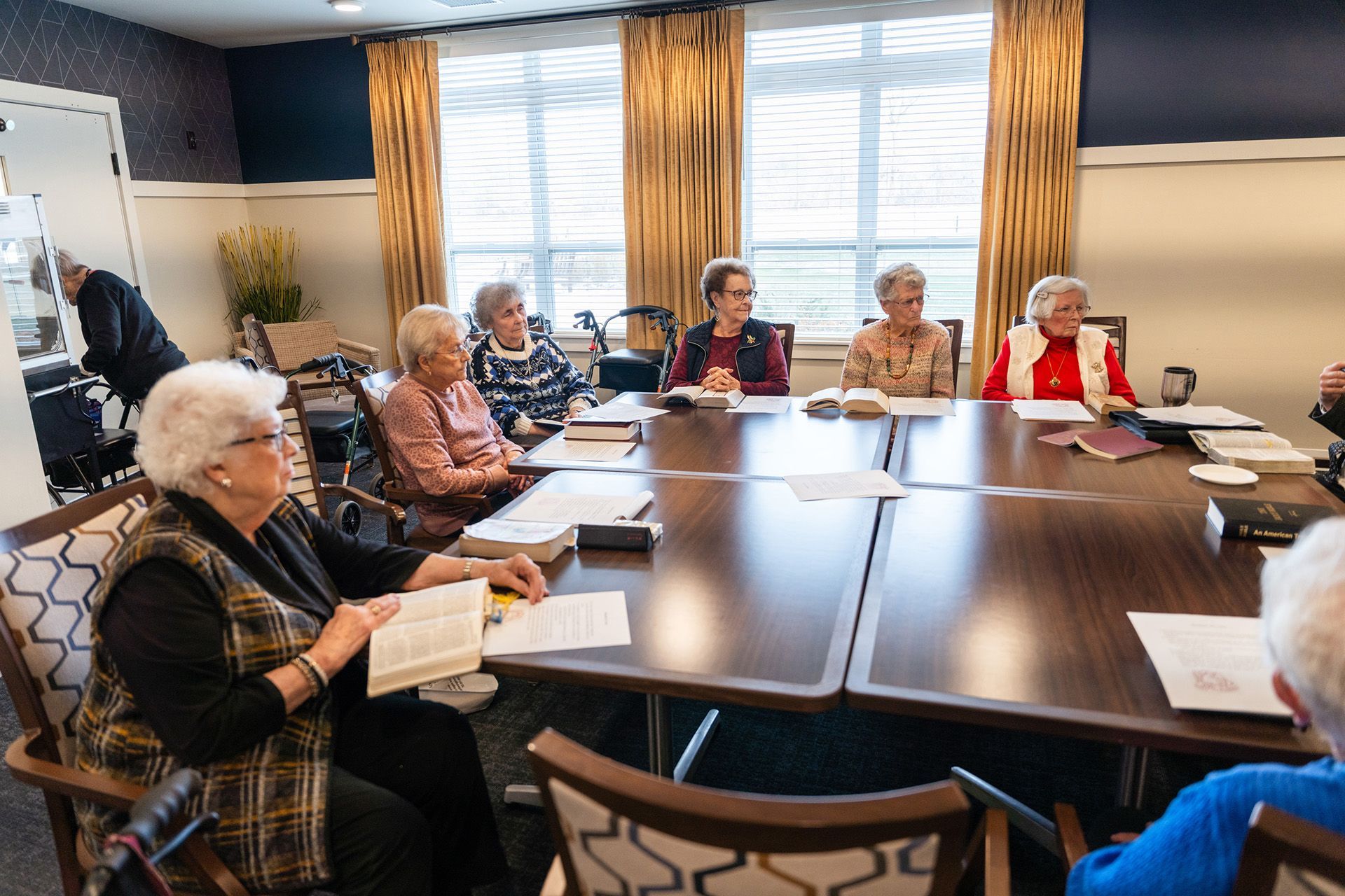 A group of people sit around a large conference table in a bright room, reading from booklets and papers.
