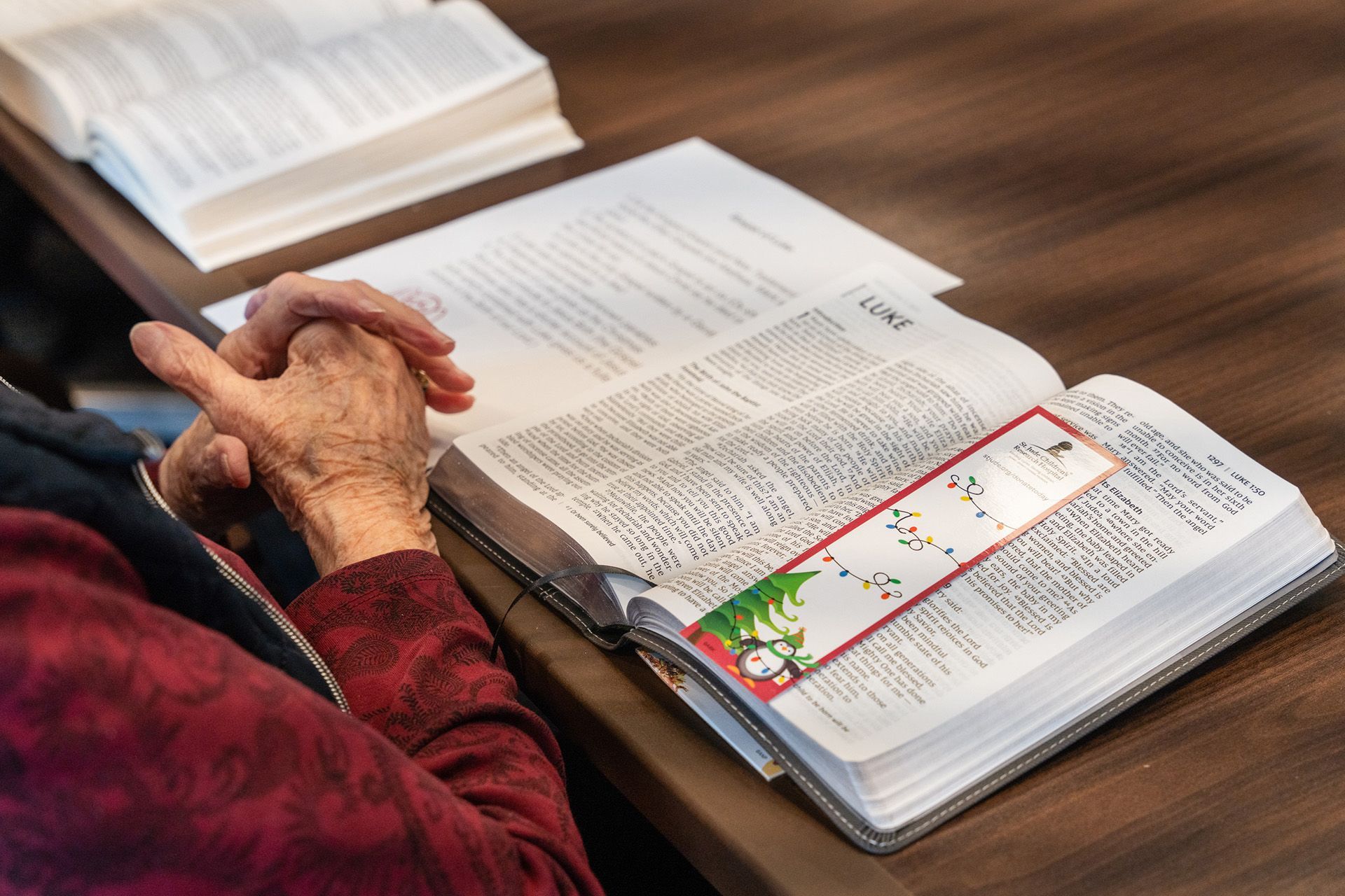 Hands clasped over an open Bible on a wooden table, with another book and papers nearby.