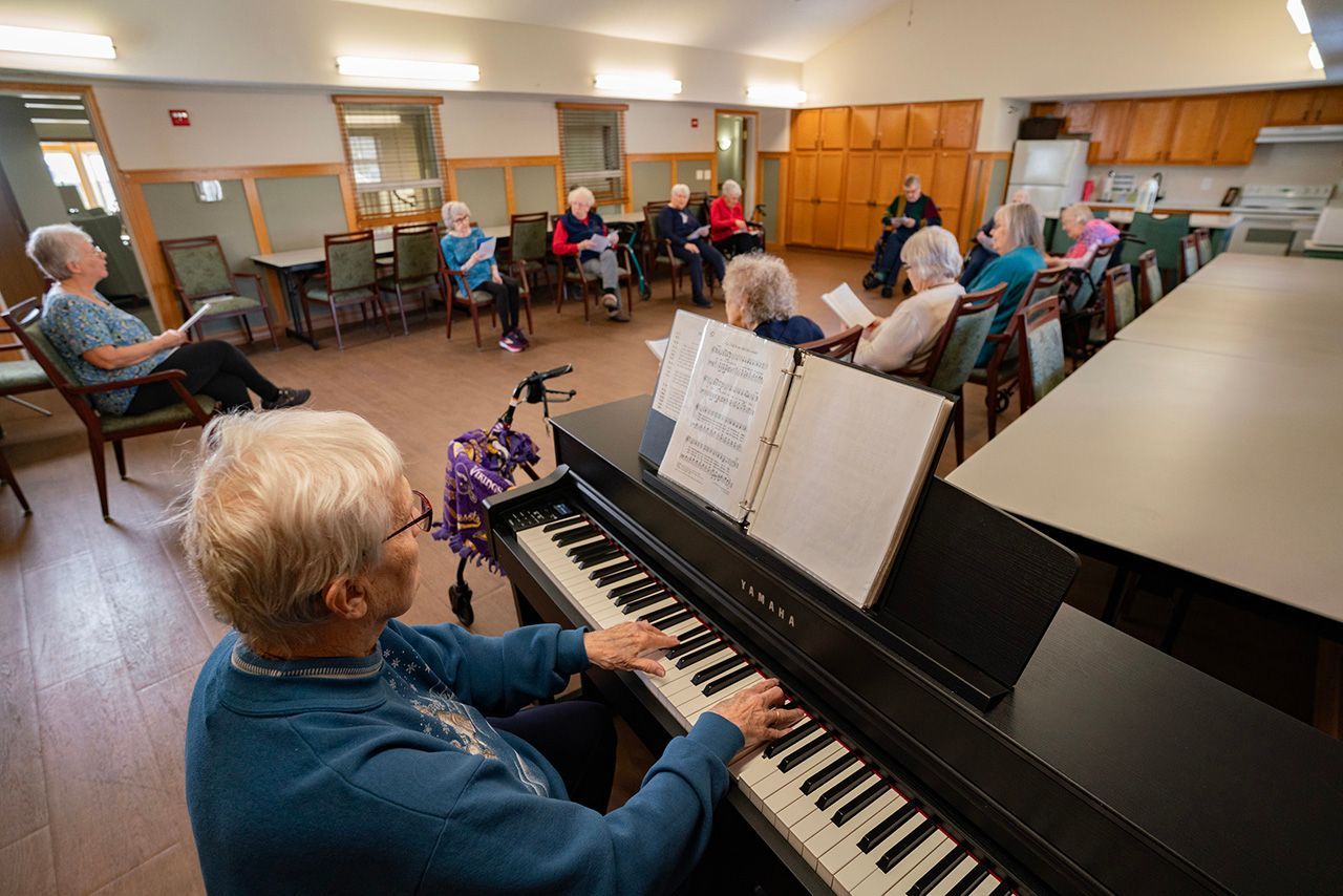 An older adult plays a piano while others sit in a circle in a community room, holding papers and appearing to sing along.