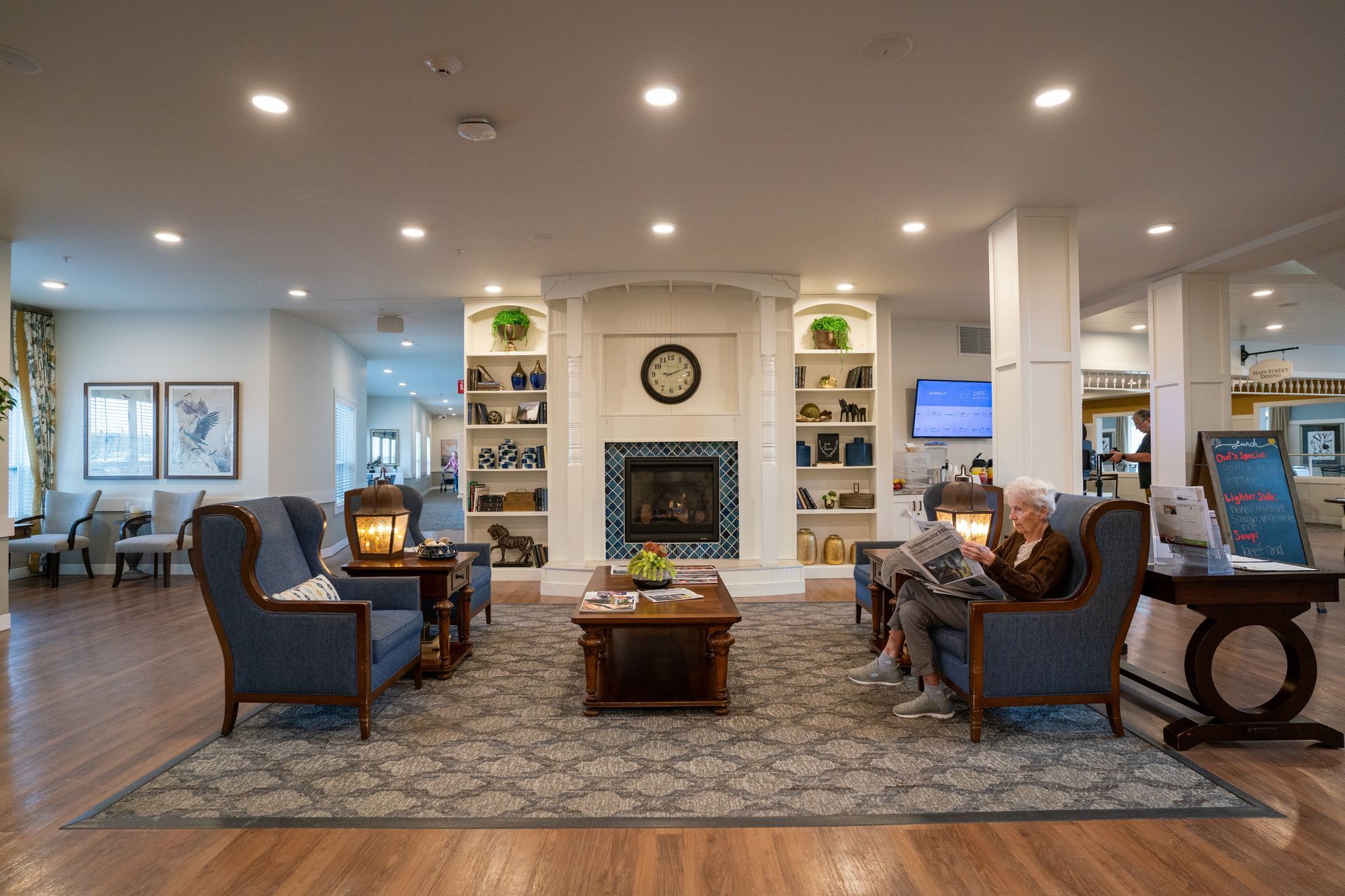 A cozy community lounge with a fireplace, bookshelves, blue armchairs, and a person reading, illuminated by recessed lights.