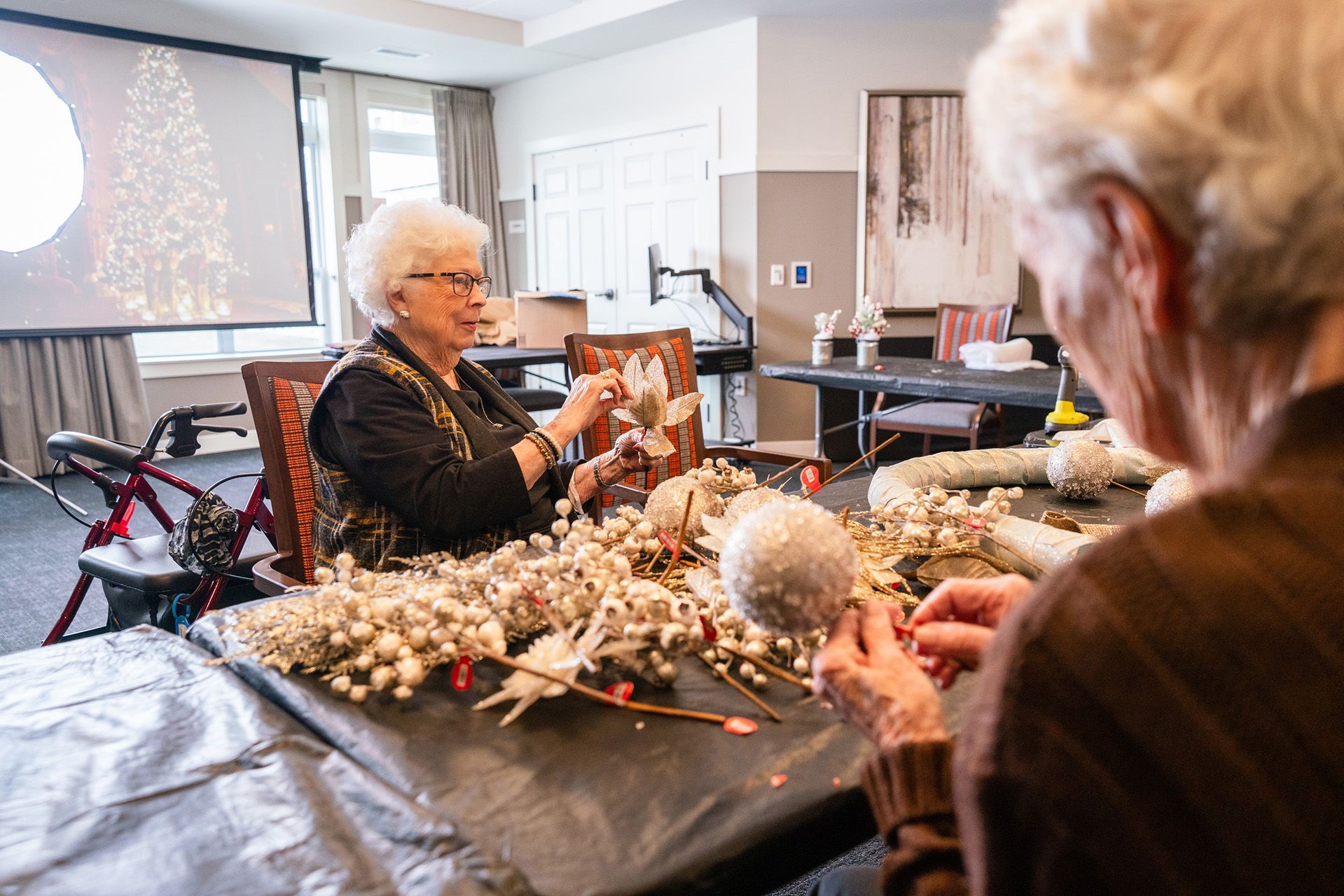 Two people work on a holiday craft project at a table in a bright room with a festive screen in the background.