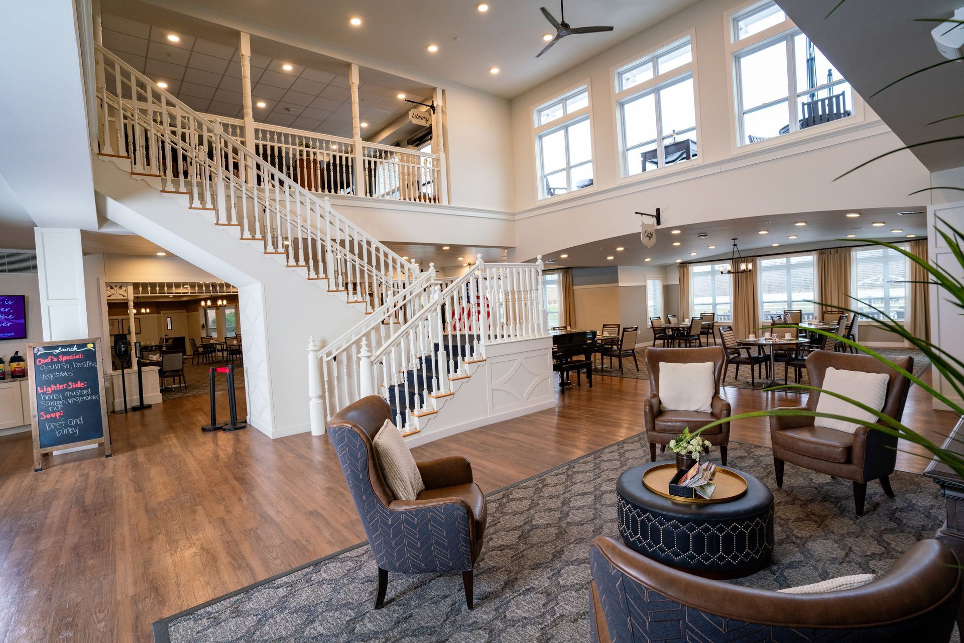 A spacious, elegant hotel lobby with a large staircase, comfortable seating area, and dining tables in the background.