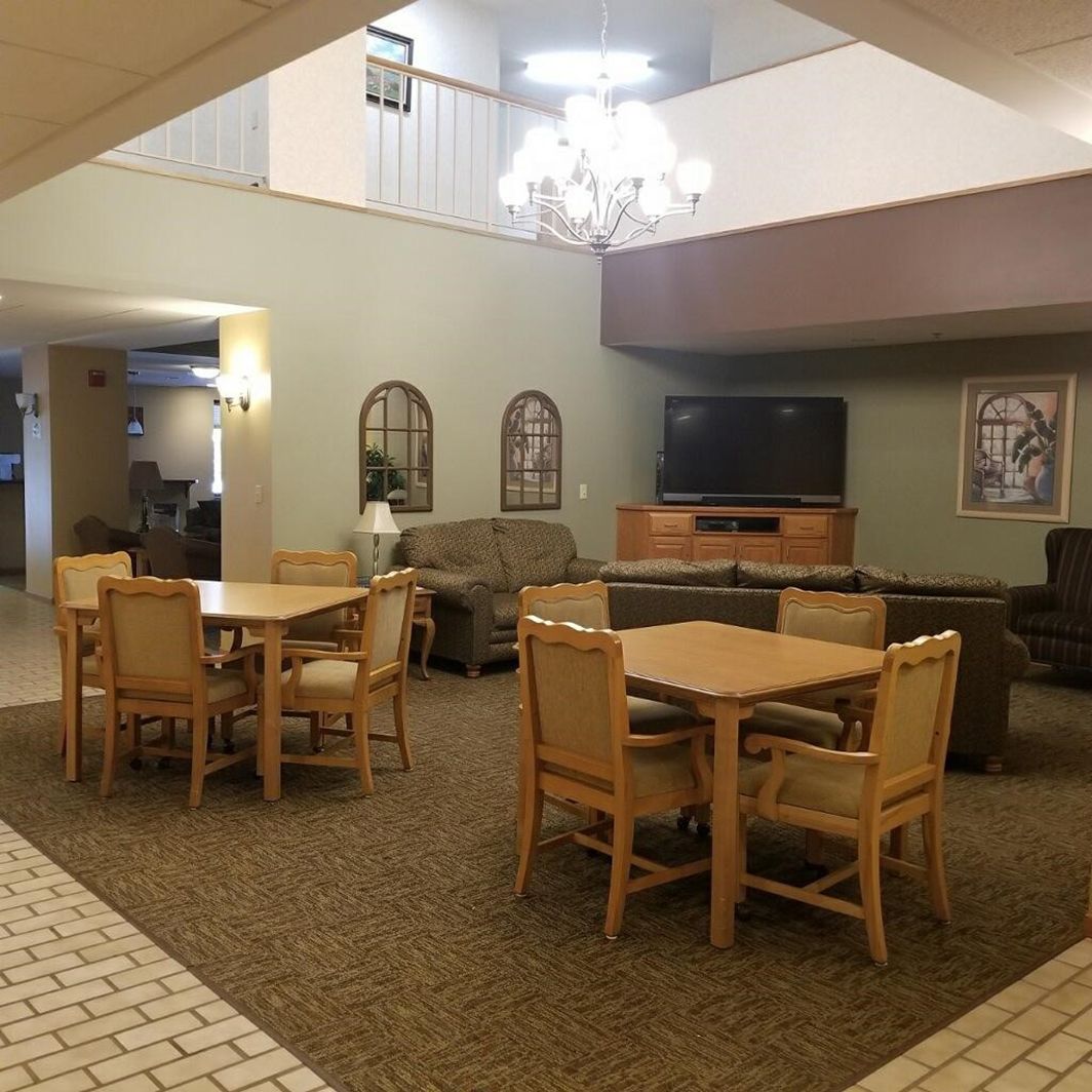 A common area with tables, chairs, sofas, a television, and a chandelier in a high-ceilinged, beige-walled room.