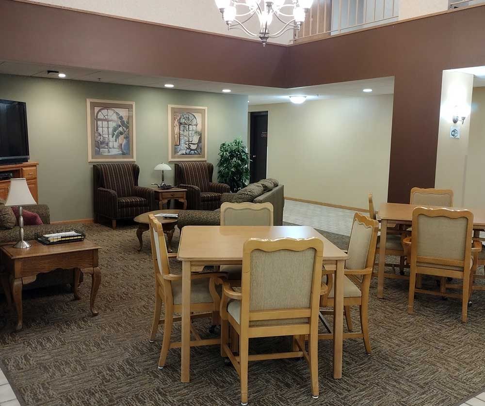 A hotel-style lounge with seating, a table, and chairs arranged in a carpeted, open-plan space with soft lighting.