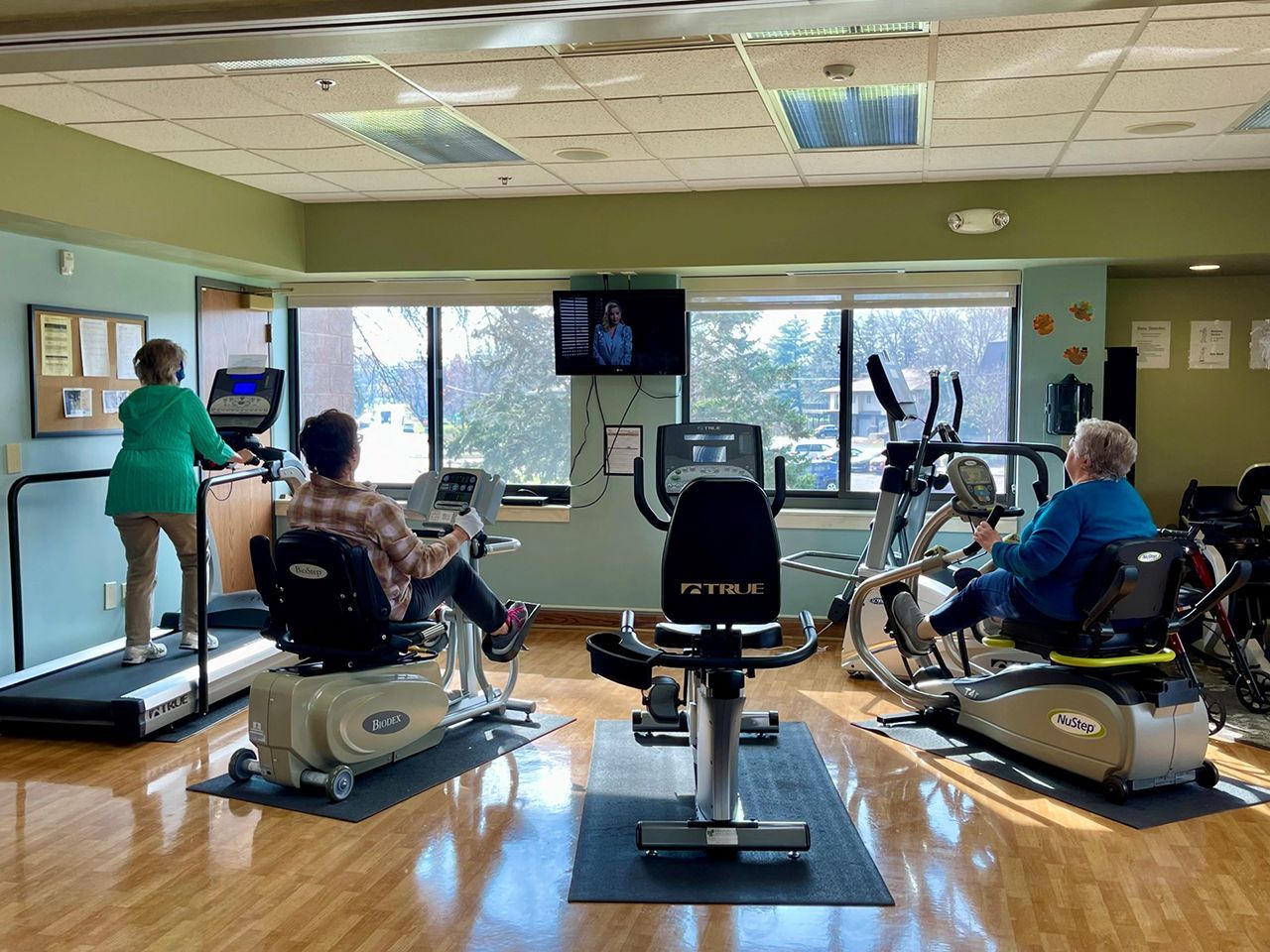 Three people exercise on different machines in a well-lit, open room with large windows and wood floors.