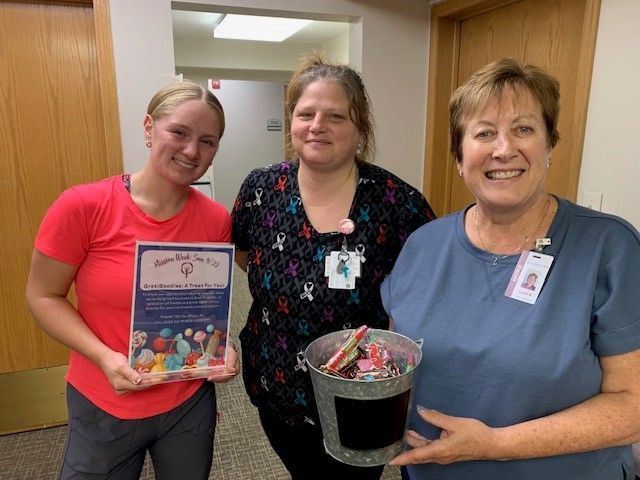 Three people smiling indoors; one holds a flyer about yarn donations, another holds a metal bucket filled with yarn balls.