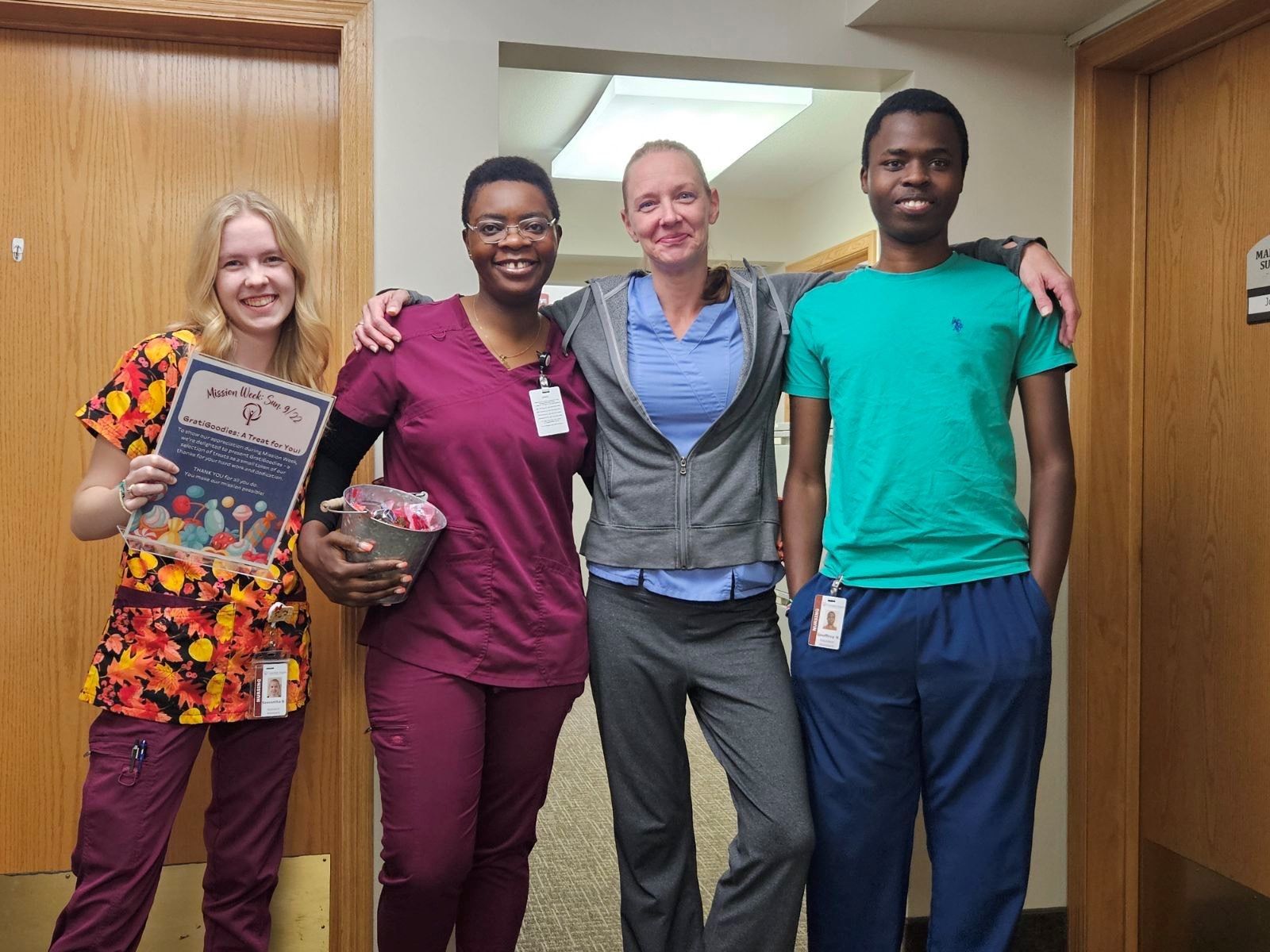 Four people standing in a hallway, smiling and posing for a photo. One person holds a card and a small gift.