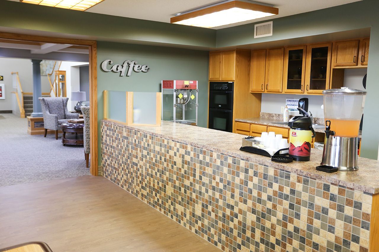 A coffee and beverage service counter with a mosaic-tiled front and wooden cabinets in a neutral-toned common area.