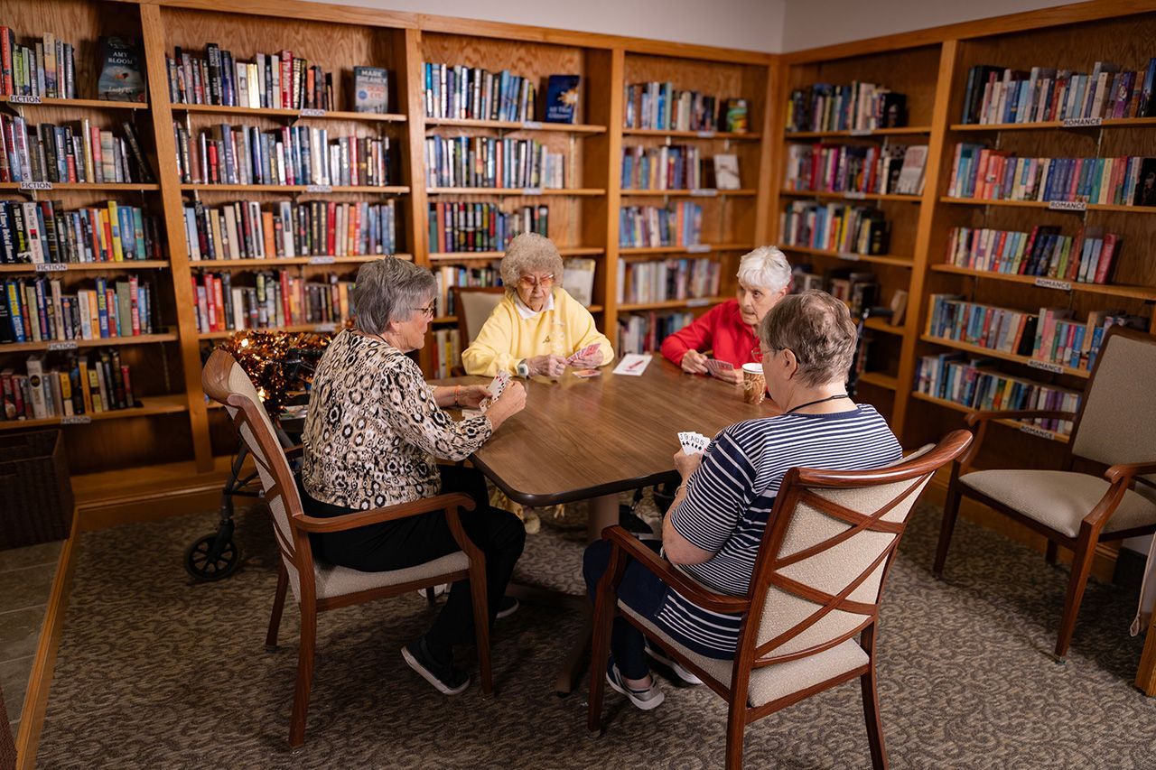 Four people sitting at a table playing cards in a library with wooden bookshelves lining the walls.