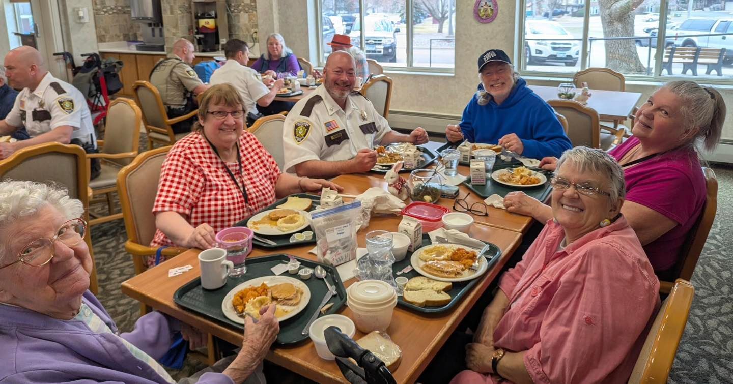 People enjoy a lunch in a bright dining hall, seated at tables with trays of food, smiling and conversing.