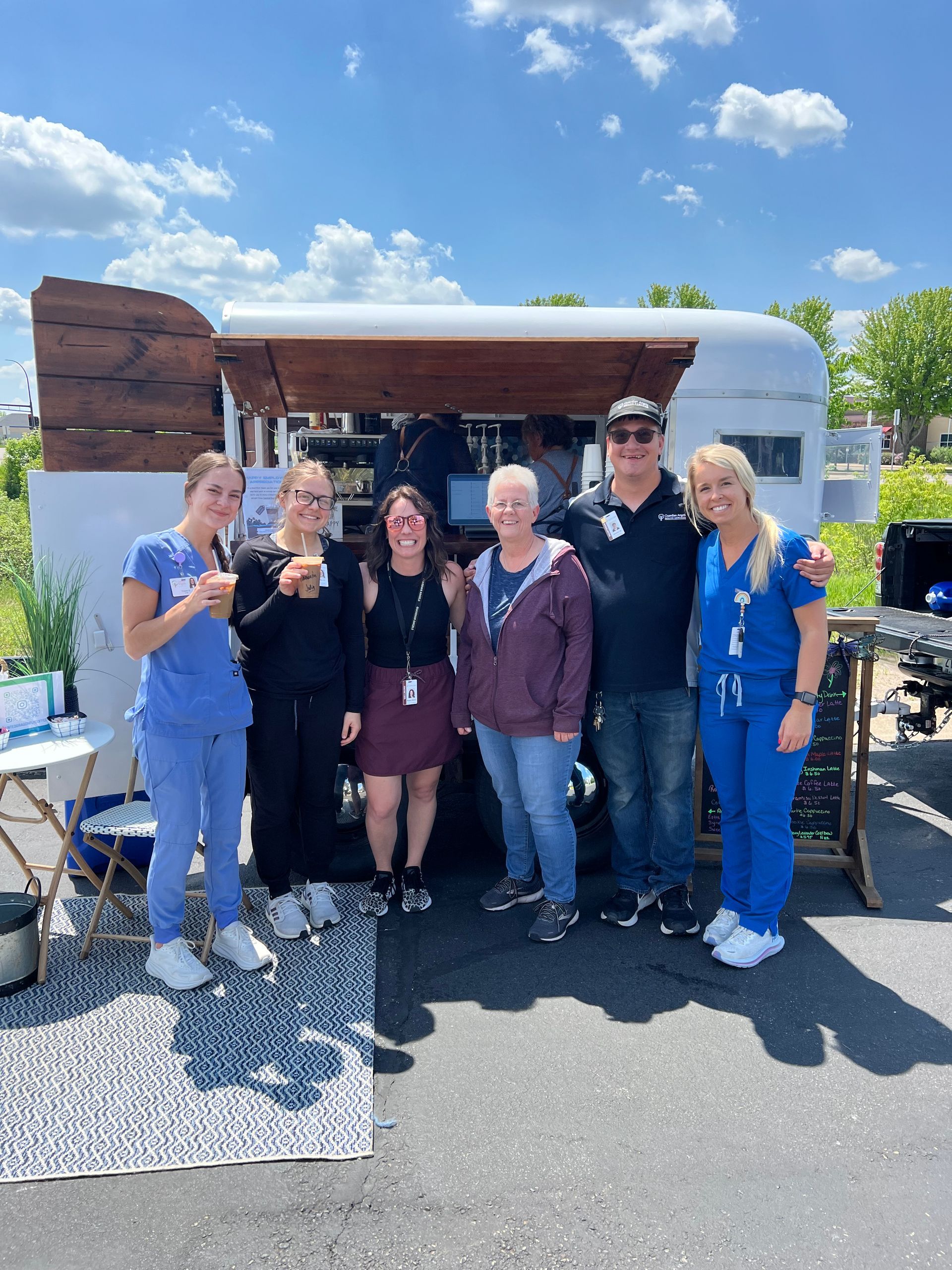 Six people stand smiling in front of a mobile food trailer on a sunny, paved lot.