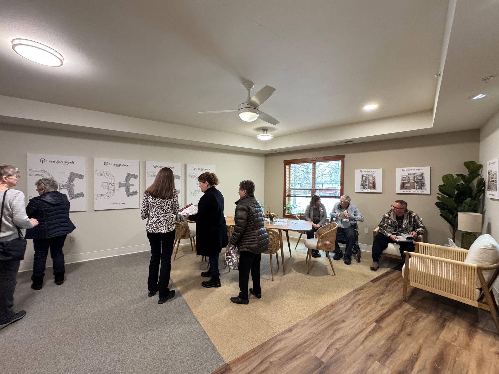 People in a brightly lit office view wall-mounted diagrams, while others sit at a table and chairs nearby.