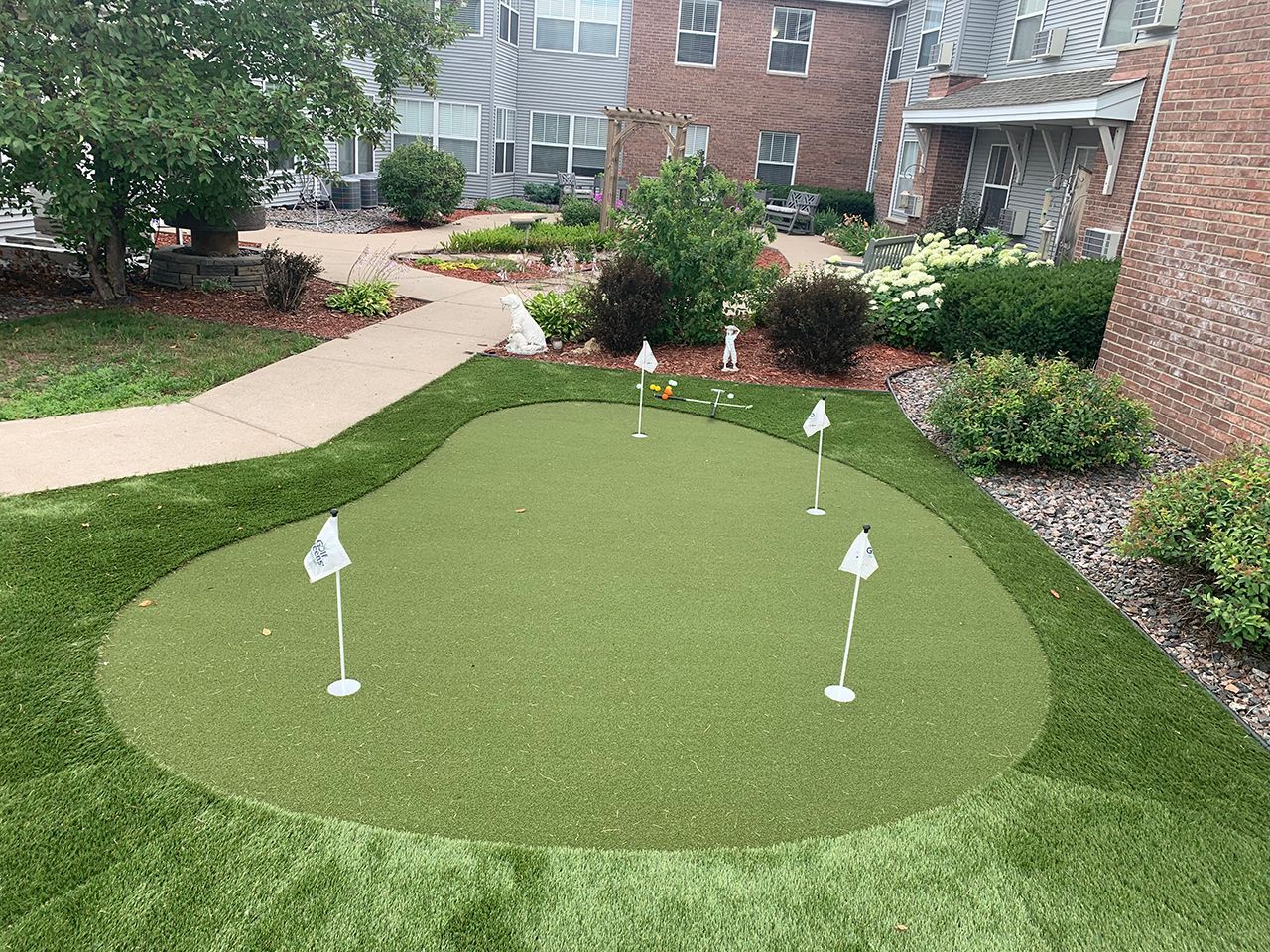 A backyard putting green with four white flags, surrounded by landscaping, a sidewalk, and a brick building.