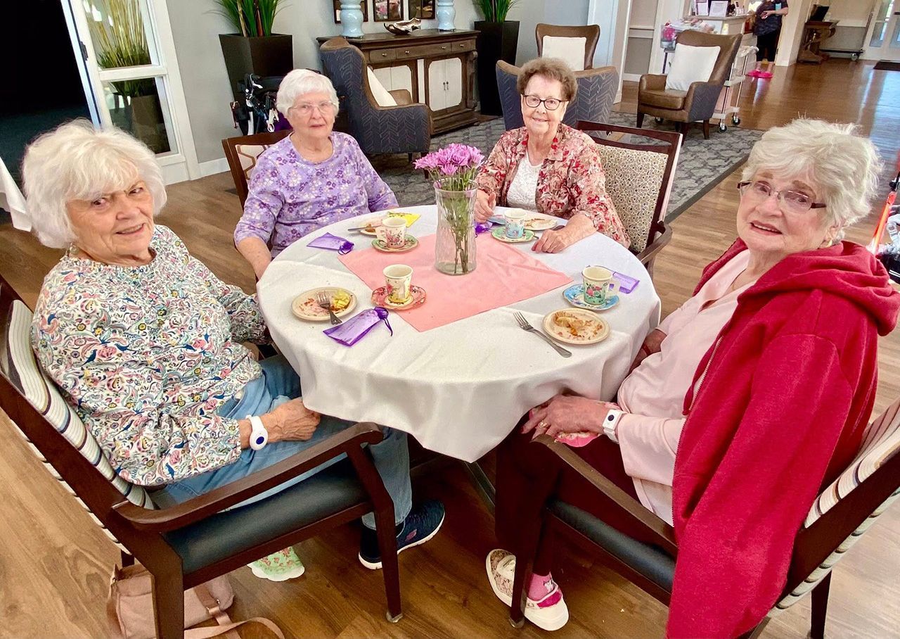 Four people sit around a round dining table with a pink cloth, small plates, and a vase of flowers in a brightly lit room.