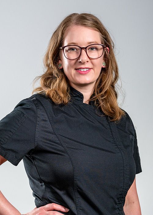 A person with shoulder-length wavy hair and glasses, wearing a black short-sleeved uniform against a light gray background.