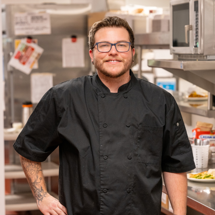 A person with glasses and arm tattoos wears a black chef’s coat while smiling in a professional kitchen.