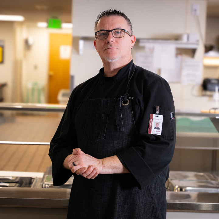 A chef in a black uniform and glasses standing in a commercial kitchen with hands clasped, wearing an identification badge.
