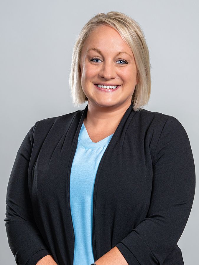 A smiling person with blonde hair wearing a light blue V-neck top under a black cardigan against a light grey background.
