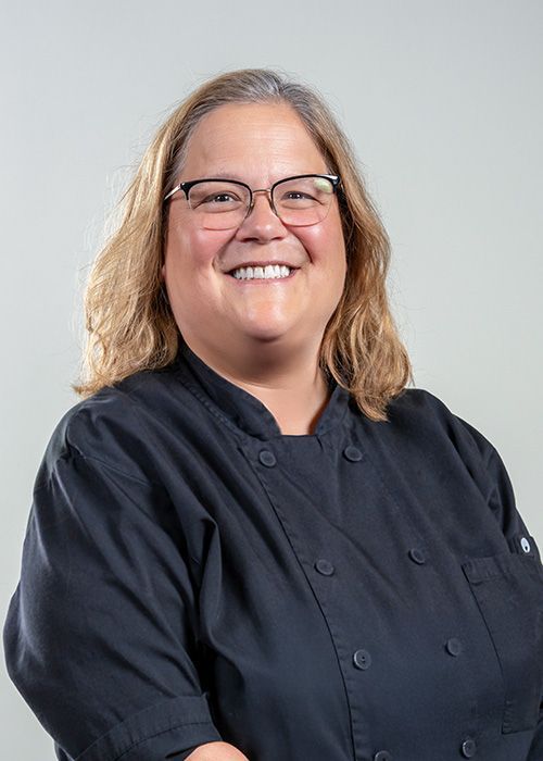 A person smiling, wearing a black chef’s coat, against a neutral gray background.