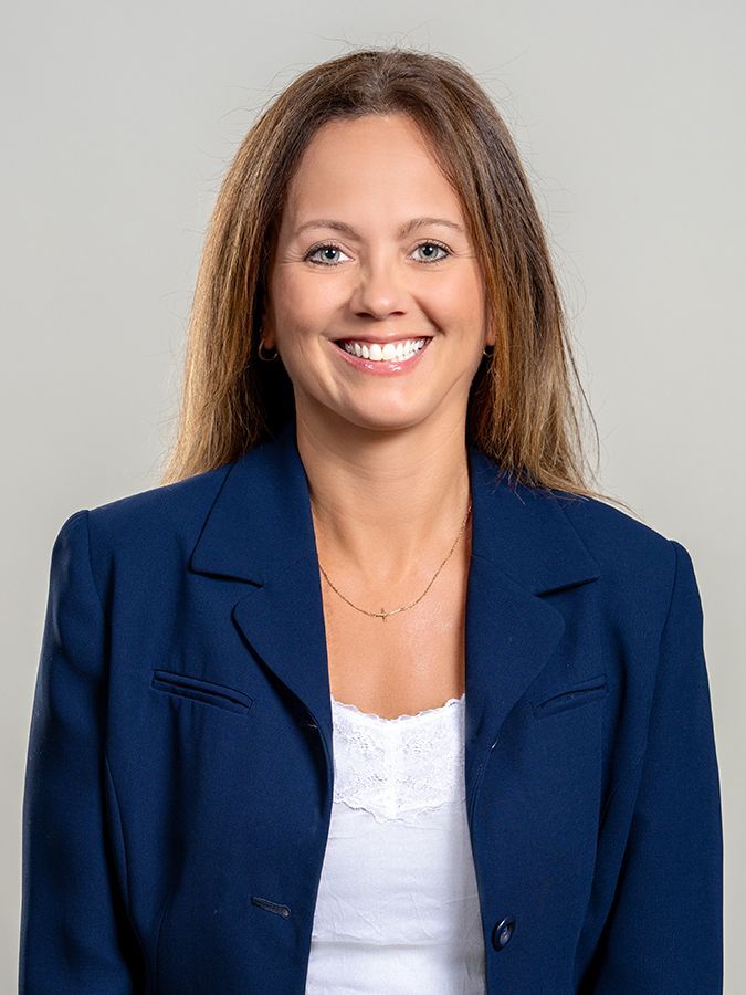 A person with shoulder-length brown hair, smiling, wearing a dark blue blazer over a white shirt against a gray background.