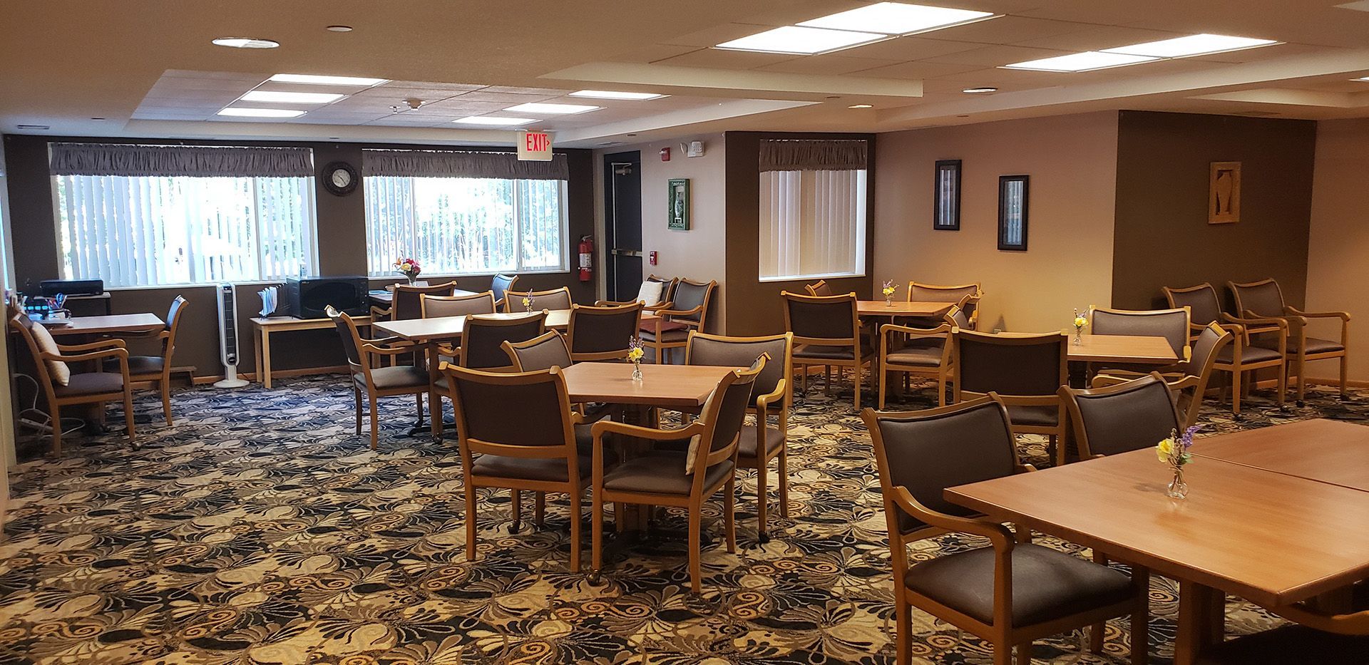 Empty dining room with wooden tables, dark chairs, and patterned carpeting, illuminated by recessed ceiling lights.