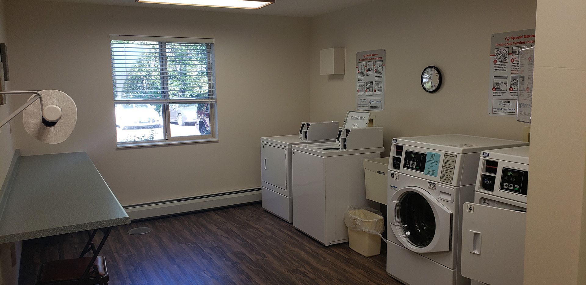 A bright, clean laundry room featuring white washers, dryers, a folding table, and informative signs on the walls.