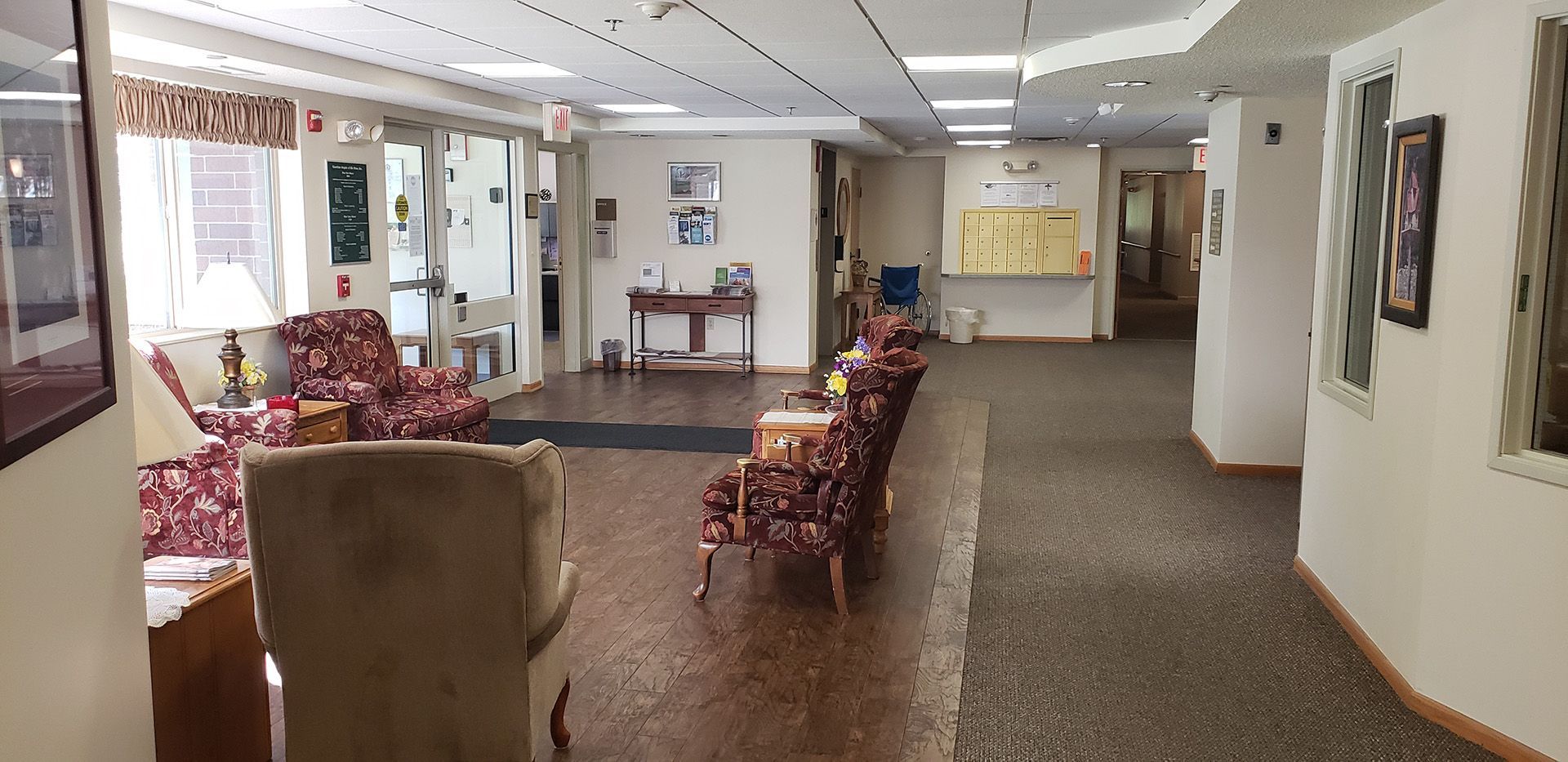 A well-lit building lobby with patterned armchairs on a hardwood floor, leading to a hallway with carpeted flooring.