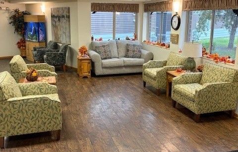 A cozy lounge area featuring patterned armchairs, a gray sofa, wood floors, and windows with fall decorations.